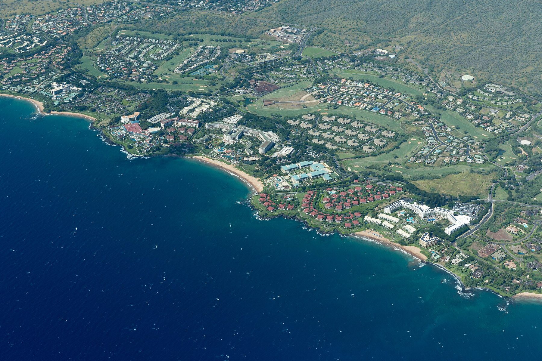 Aerial view of a coastline with several large resort hotels, pools, red-roofed condominiums, golf fairways and housing behind crescent sand beaches and dark blue sea.