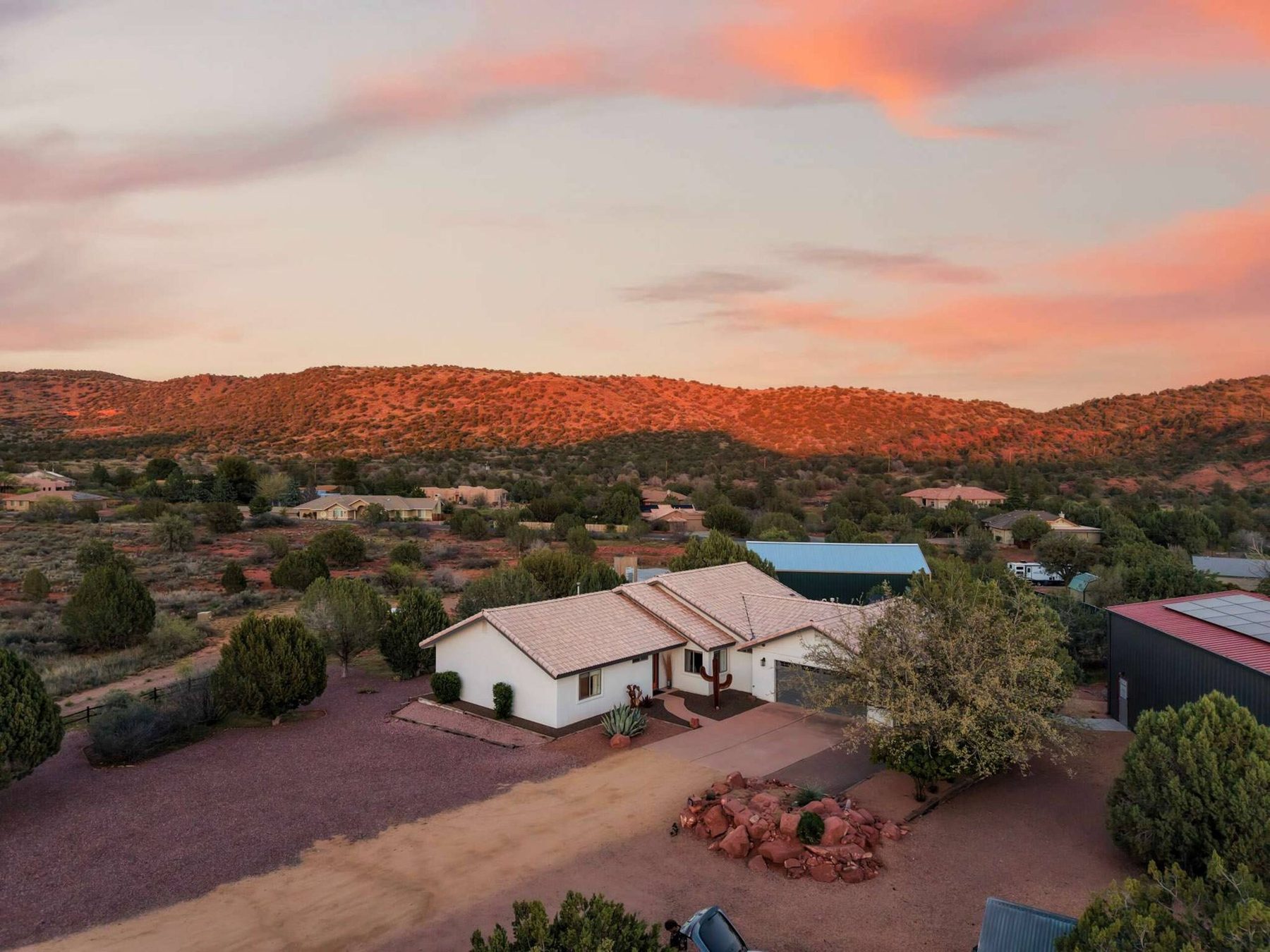 Drone view of a white single-storey house with a pale tile roof, a gravel yard and juniper trees under pink sunset clouds; low hills glow orange behind, and a dark metal building with roof panels sits at the right edge.