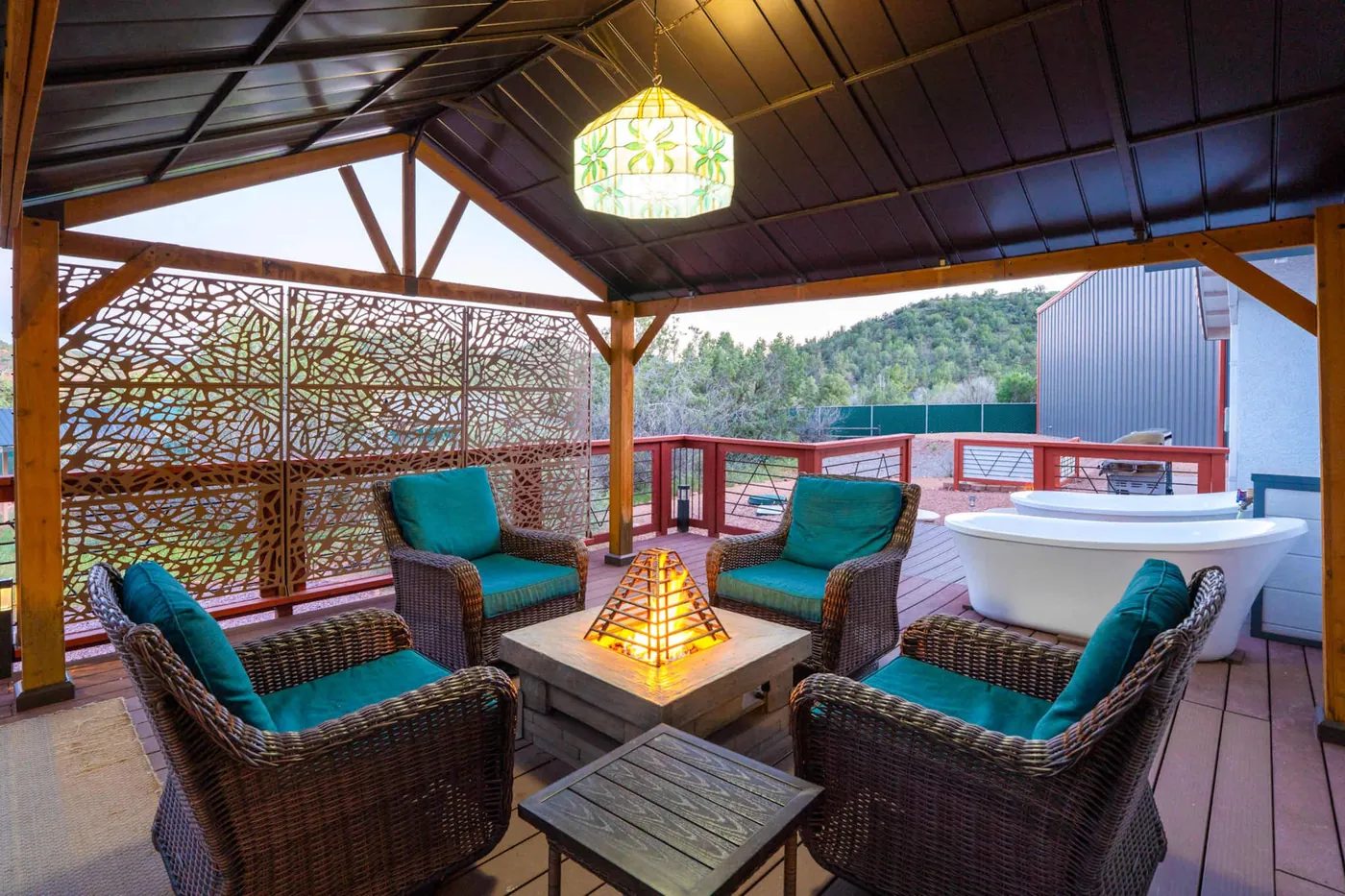 Four wicker armchairs with teal cushions surround a lit gas fire table under a timber pergola with a stained-glass pendant lamp; two white freestanding tubs and the wall of a grey metal building are visible behind.