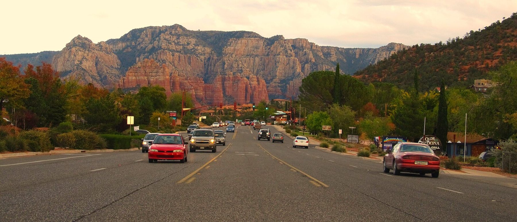 A wide multi-lane road with cars travelling in both directions and traffic lights ahead, lined with trees and roadside signs; tall layered red and cream cliffs fill the background under a pale sky.