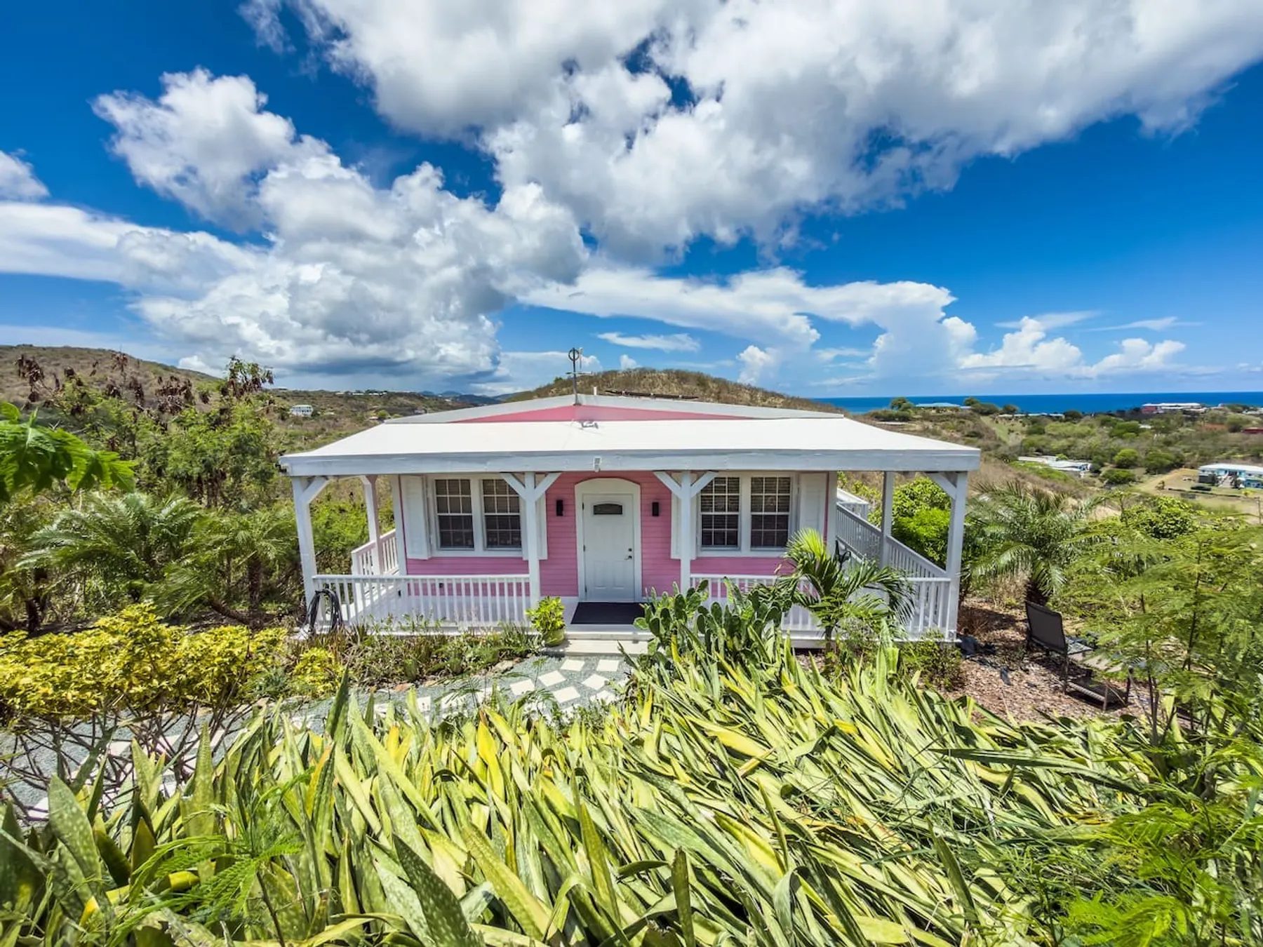 A small pink wooden cottage with white trim and a wrap-around porch, on a hillside planted with agave, palms and cactus, under large white clouds with the sea in the distance.