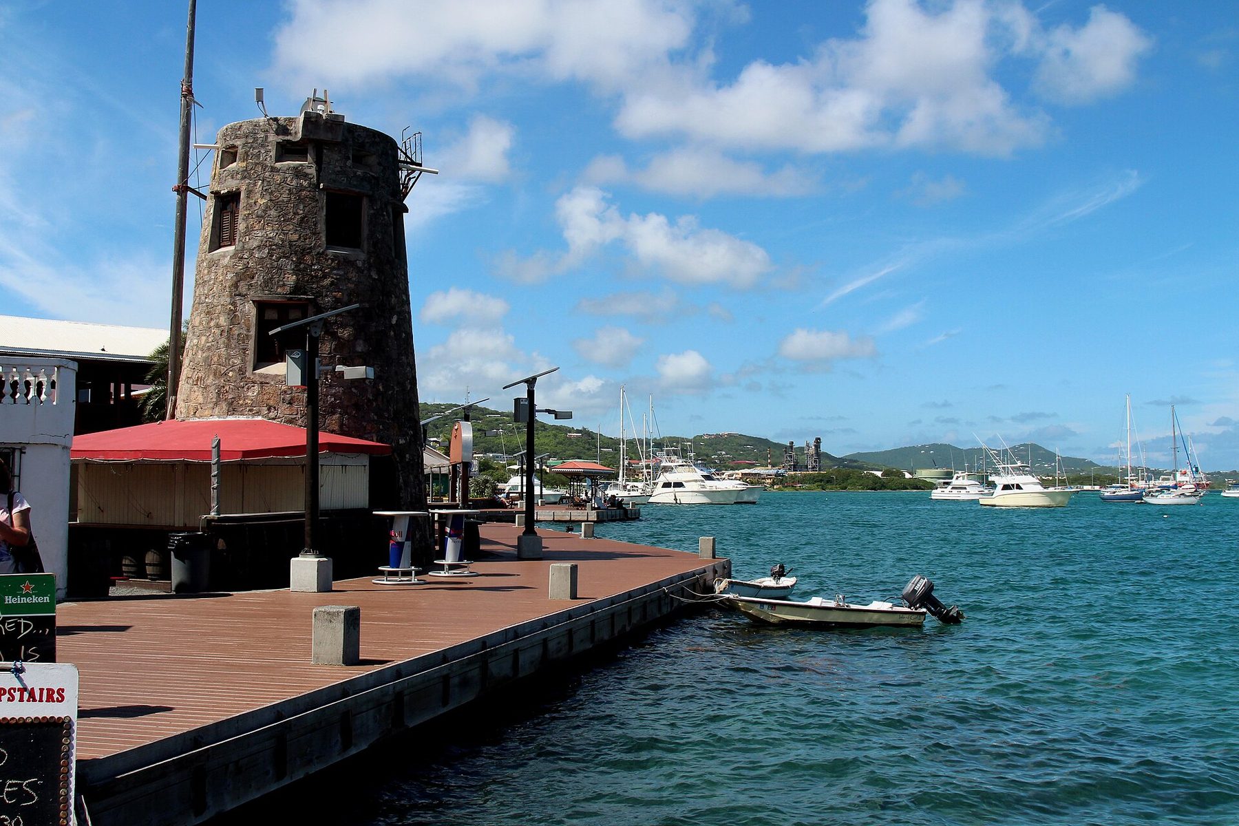 A wooden boardwalk beside a round stone tower with a red awning at its base, with small boats tied alongside and yachts moored in a harbour under a blue sky.