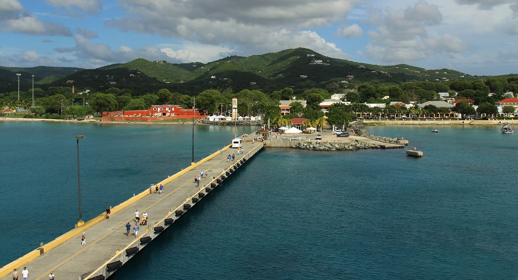 A long concrete pier with people walking along it, leading to a small town with a red fort and a clock tower on the shore, backed by green hills.