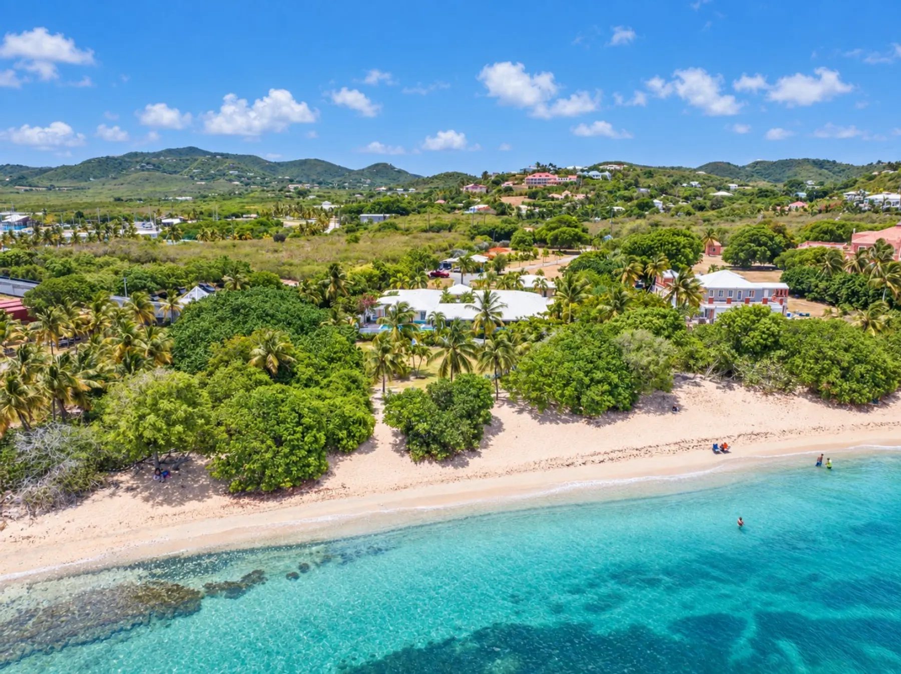 An aerial view of a pale sand beach and clear turquoise shallows, with a low white-roofed house set among palms and sea grape trees behind the sand and green hills beyond.