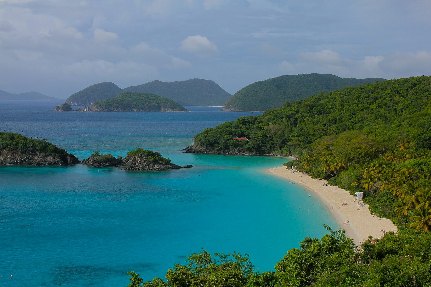 A crescent of white sand lined with palms beside clear turquoise water, with small rocky islets offshore and forested hills and islands behind, seen from above.
