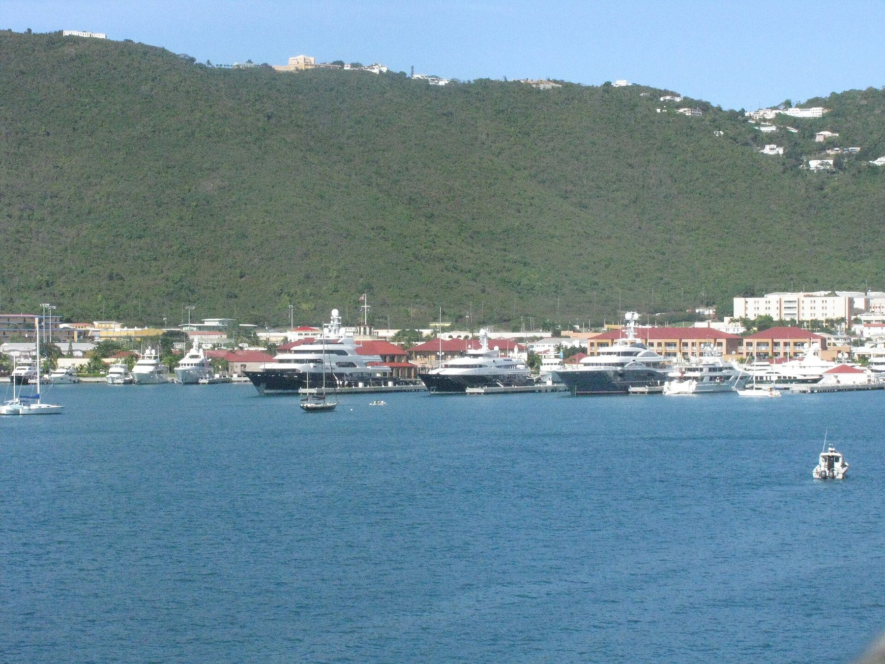 Large motor yachts moored at a marina with red-roofed buildings along the waterfront, below a steep green hillside dotted with houses, seen across blue harbour water.