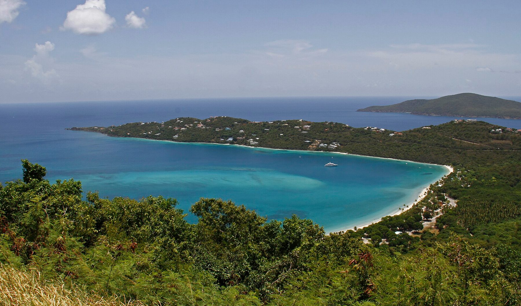 A wide turquoise bay enclosed by a wooded peninsula with scattered houses, a long curve of white sand at its head and a single yacht at anchor, seen from a hilltop.