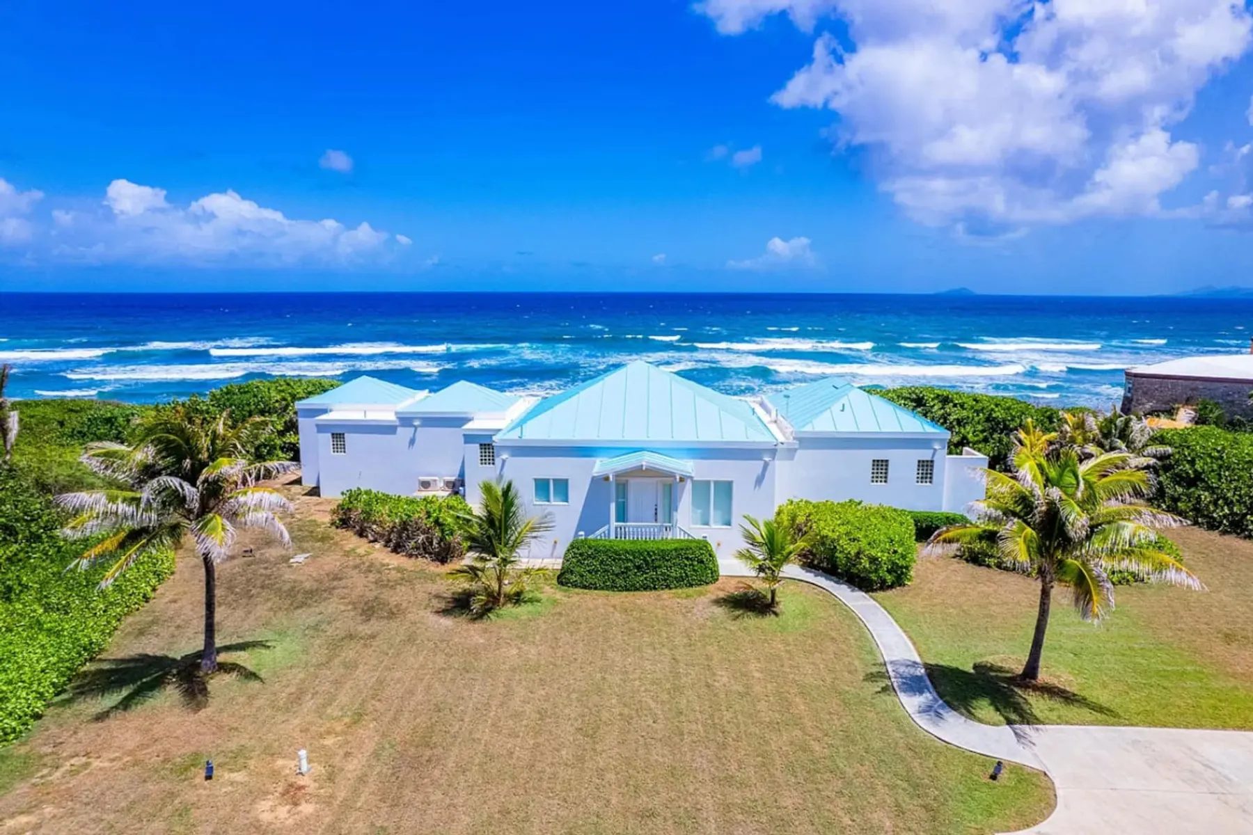 An aerial view of a single-storey white house with pale blue metal roofs, a lawn with young palms in front and the open sea with lines of breaking waves directly behind it.