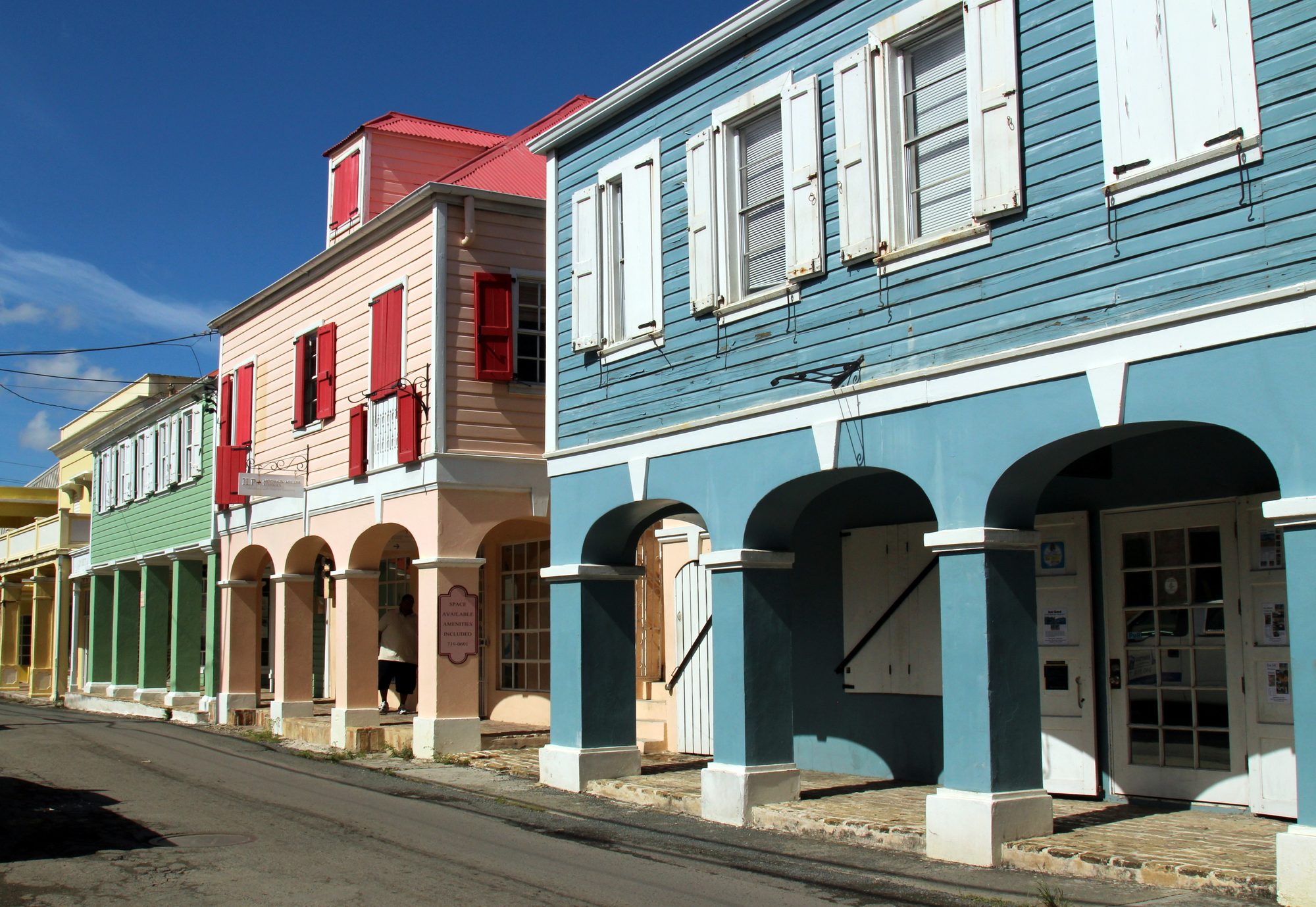 Two-storey buildings with shuttered upper floors and arcaded ground floors, painted blue, pink, green and yellow, along a street in Christiansted on St. Croix