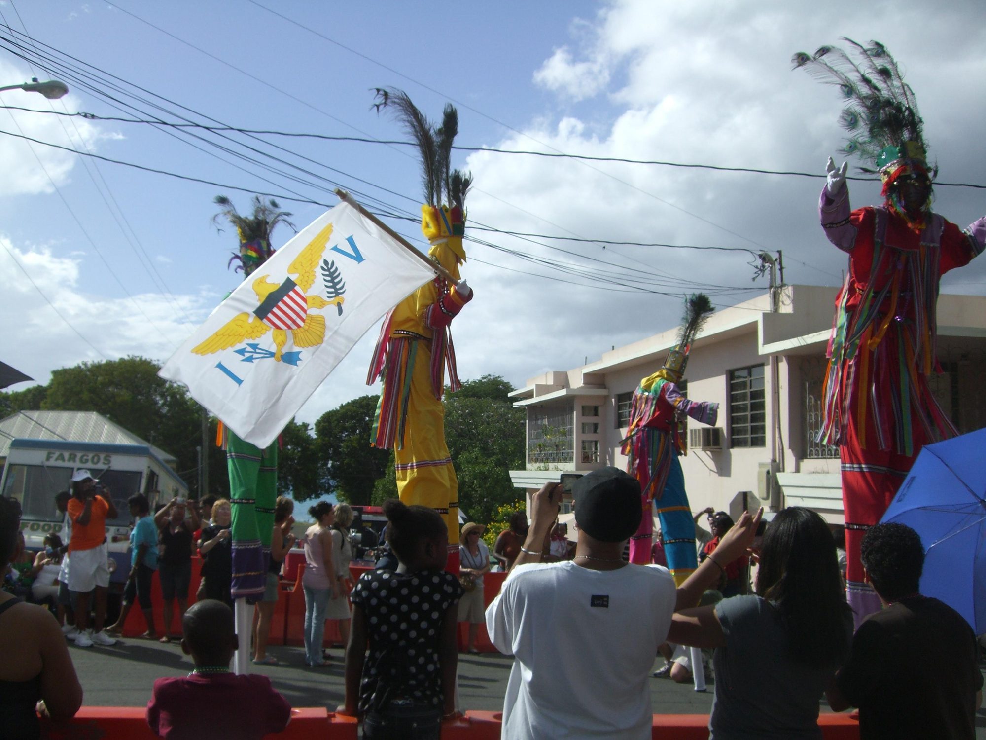 Moko jumbie stilt dancers in bright costumes, one carrying the flag of the U.S. Virgin Islands, performing above a crowd at a street parade on St. Croix