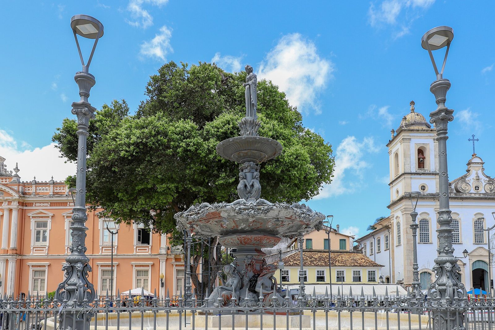 An ornate grey metal fountain topped by a statue on the Terreiro de Jesus, behind iron railings between tall lamp posts, with a salmon-pink building on the left and a white church with a bell tower on the right