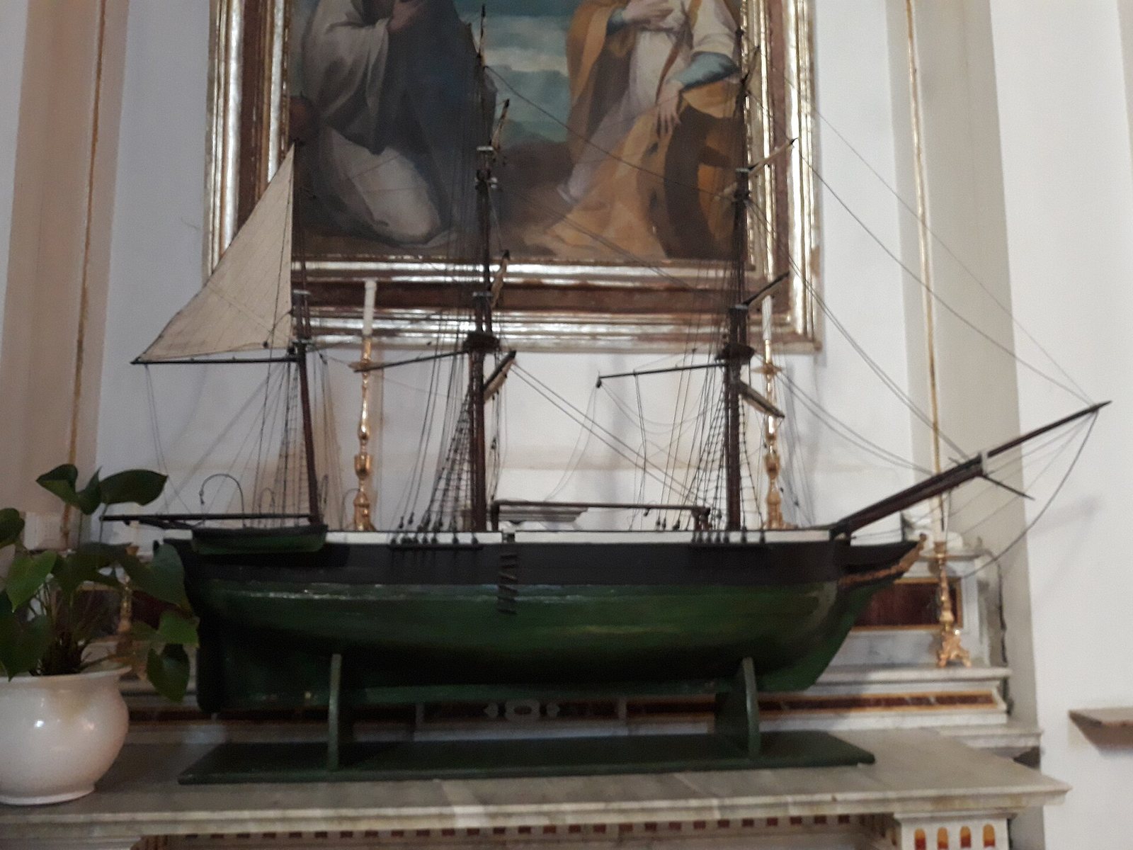 A detailed model of a sailing ship with a dark green hull, white sails, and intricate rigging stands on a shelf in front of a framed painting.