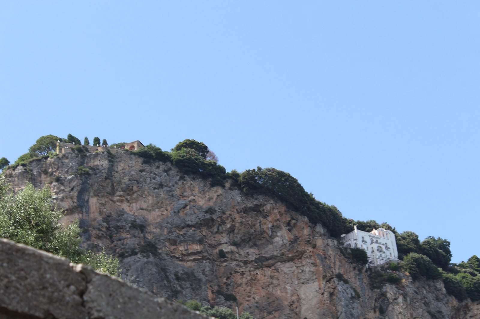 A cliff face under a clear sky, with the Terrazzo dell'lnfinito visible on the left and the white La Rondinaia villa on the right.