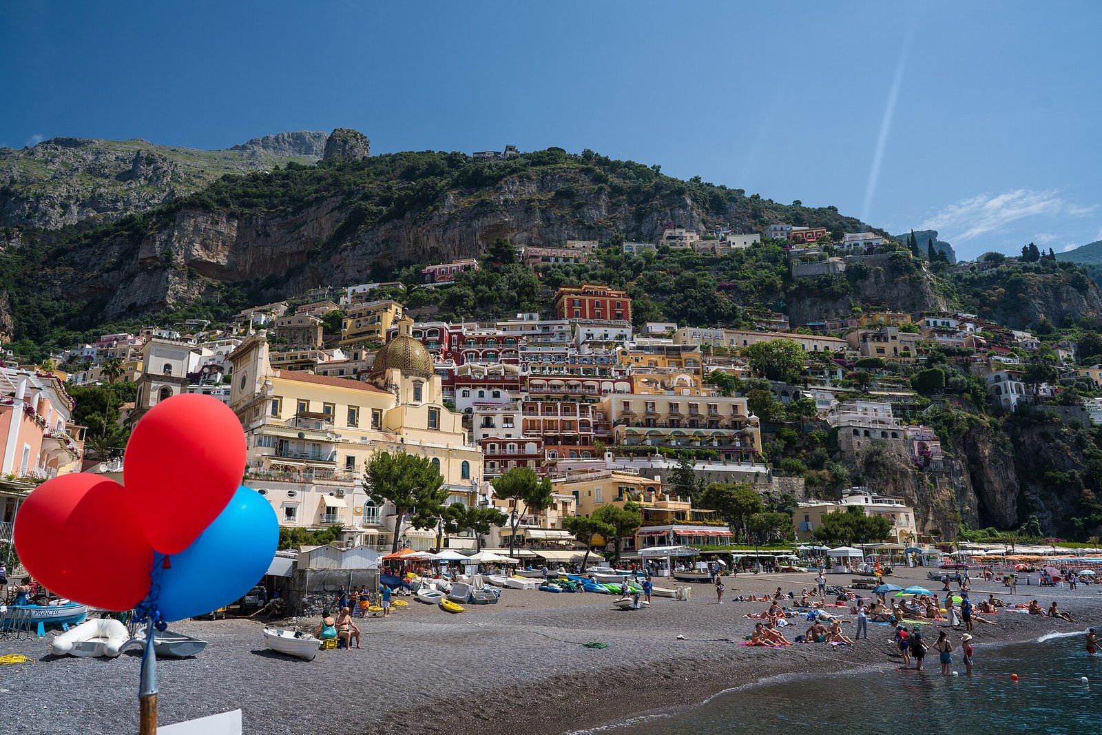 Three large balloons, two red and one blue, are on a pole at a pebbly beach with many people, boats, and umbrellas. A town climbs a steep hillside under a clear blue sky.