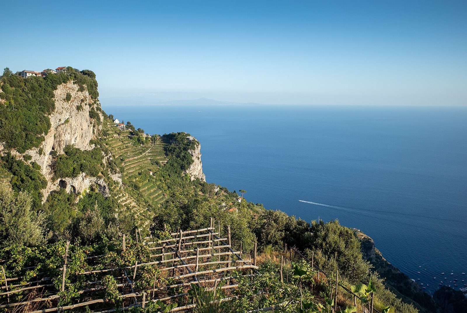 A view from above shows terraced plantations on a steep, green cliffside overlooking the deep blue sea, with a boat leaving a white wake.