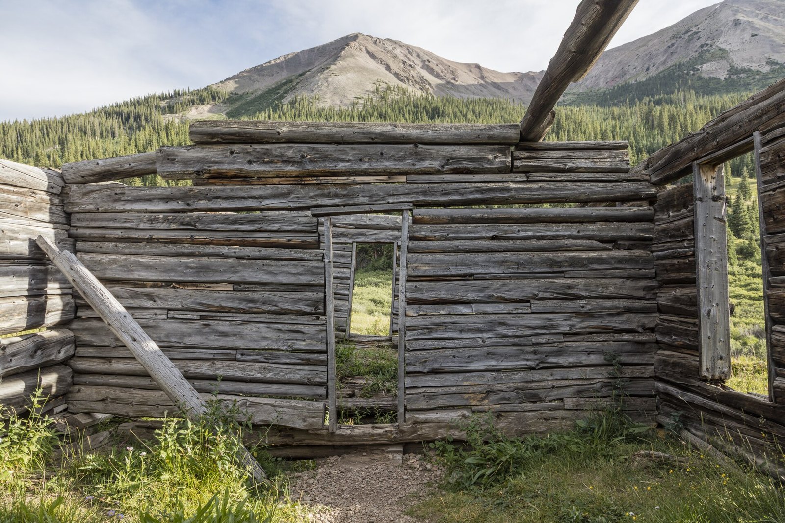 The weathered log walls of a ruined cabin with an empty doorway, in a grassy mountain valley with a peak behind
