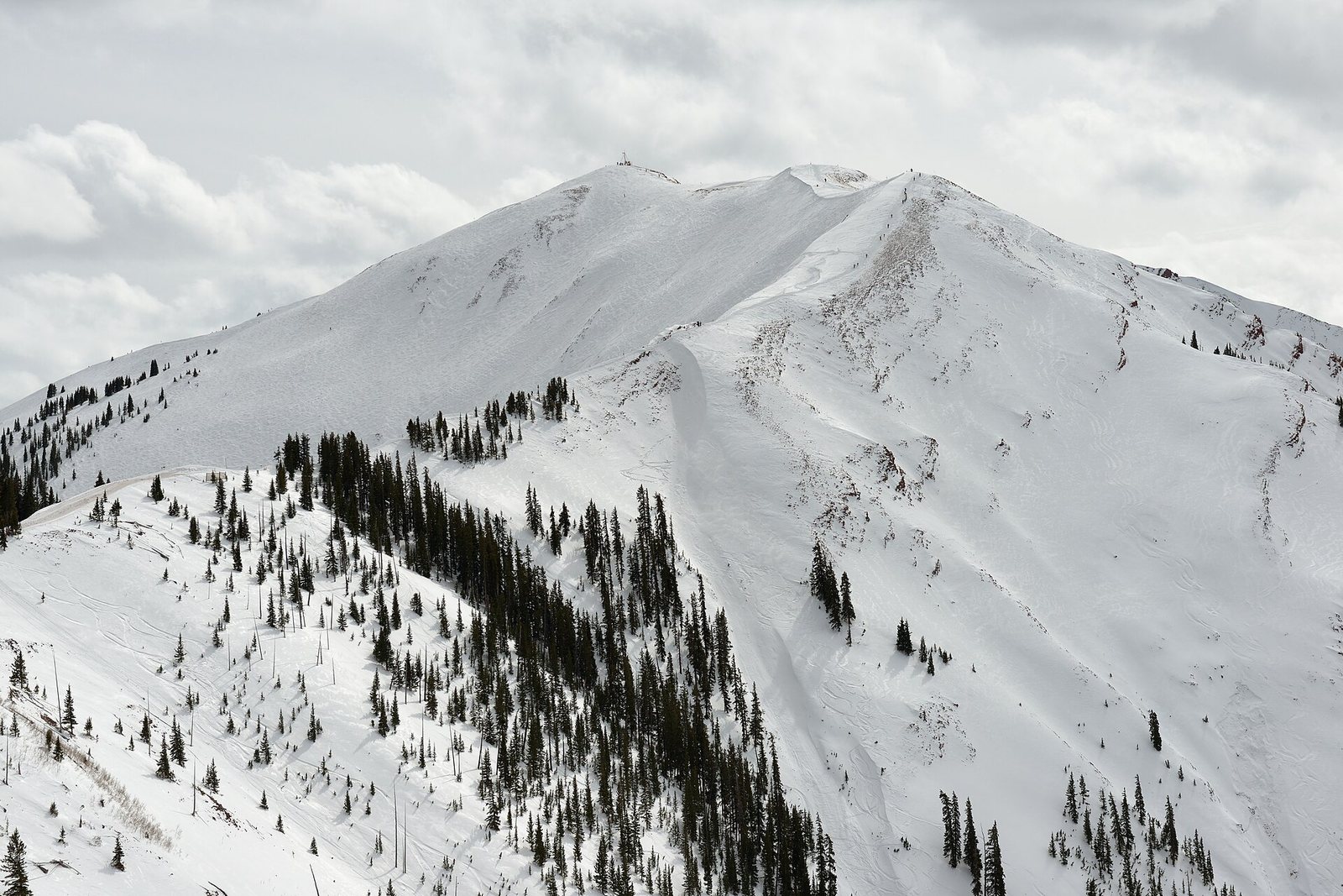 A snow-covered mountain with ski runs and scattered evergreen trees under a cloudy sky, with several people visible on the slopes.