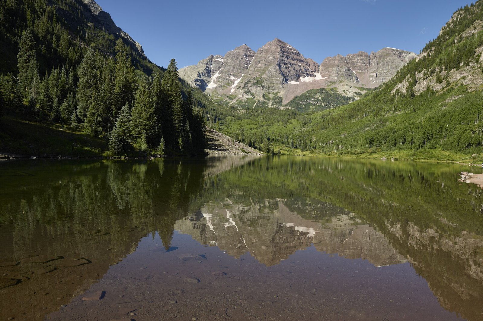The twin peaks of the Maroon Bells reflected in the still water of Maroon Lake, with dark forest on both slopes