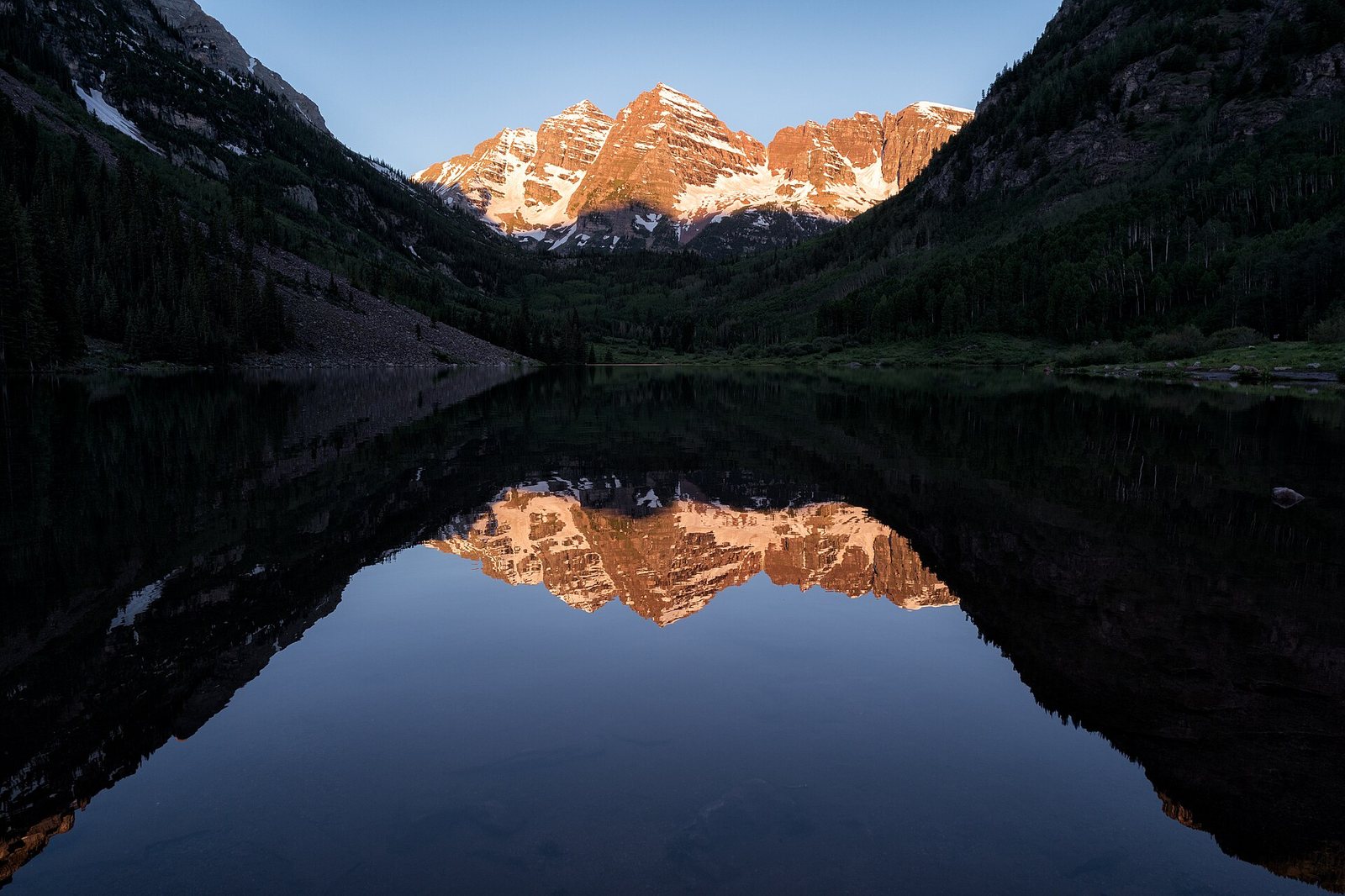 Snow-capped mountains with sunlit peaks are reflected in a still lake, flanked by dark forested slopes under a clear sky at sunrise.