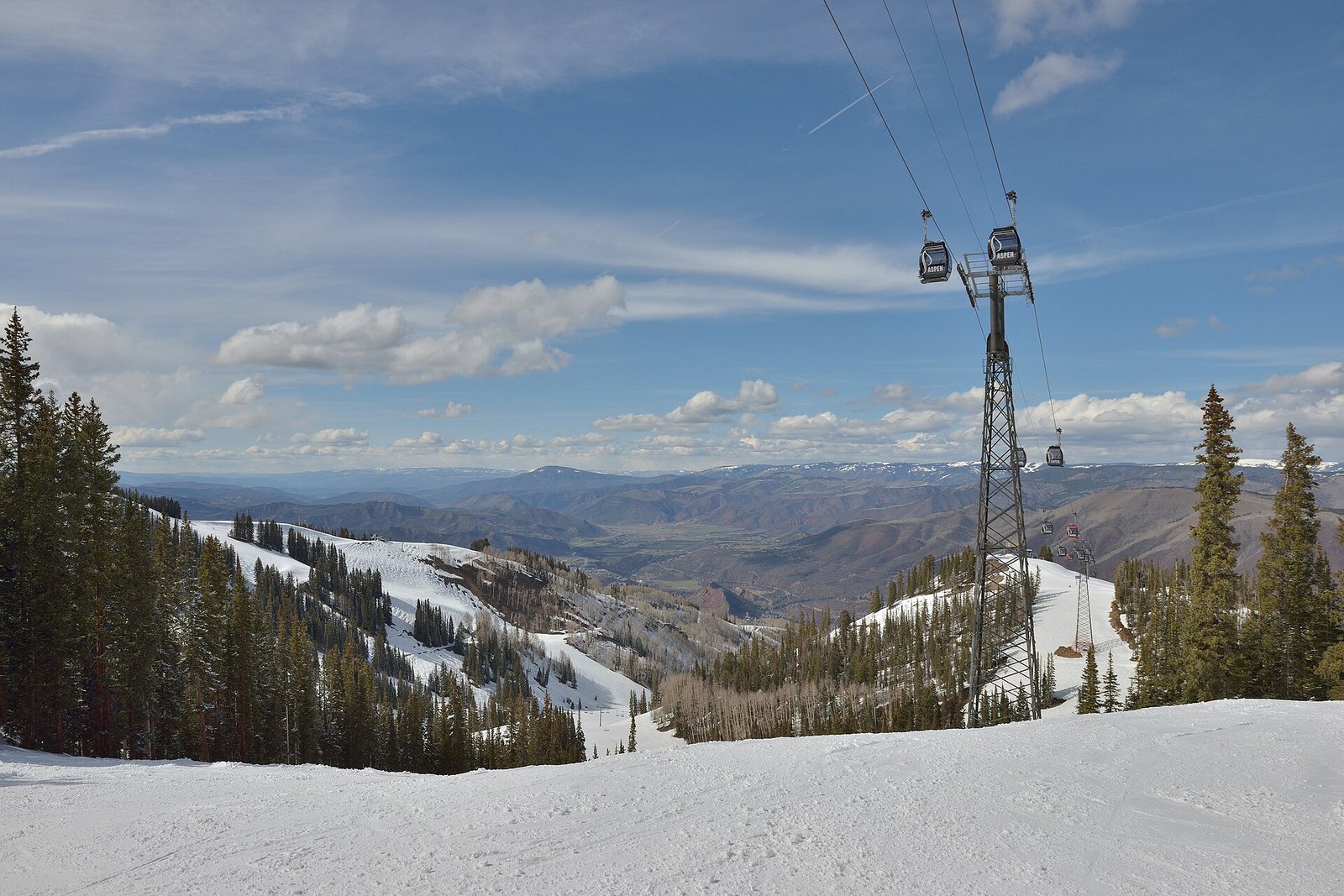 A gondola lift with several cars ascends a snowy mountain slope lined with pine trees, with a valley and distant mountains under a cloudy sky.