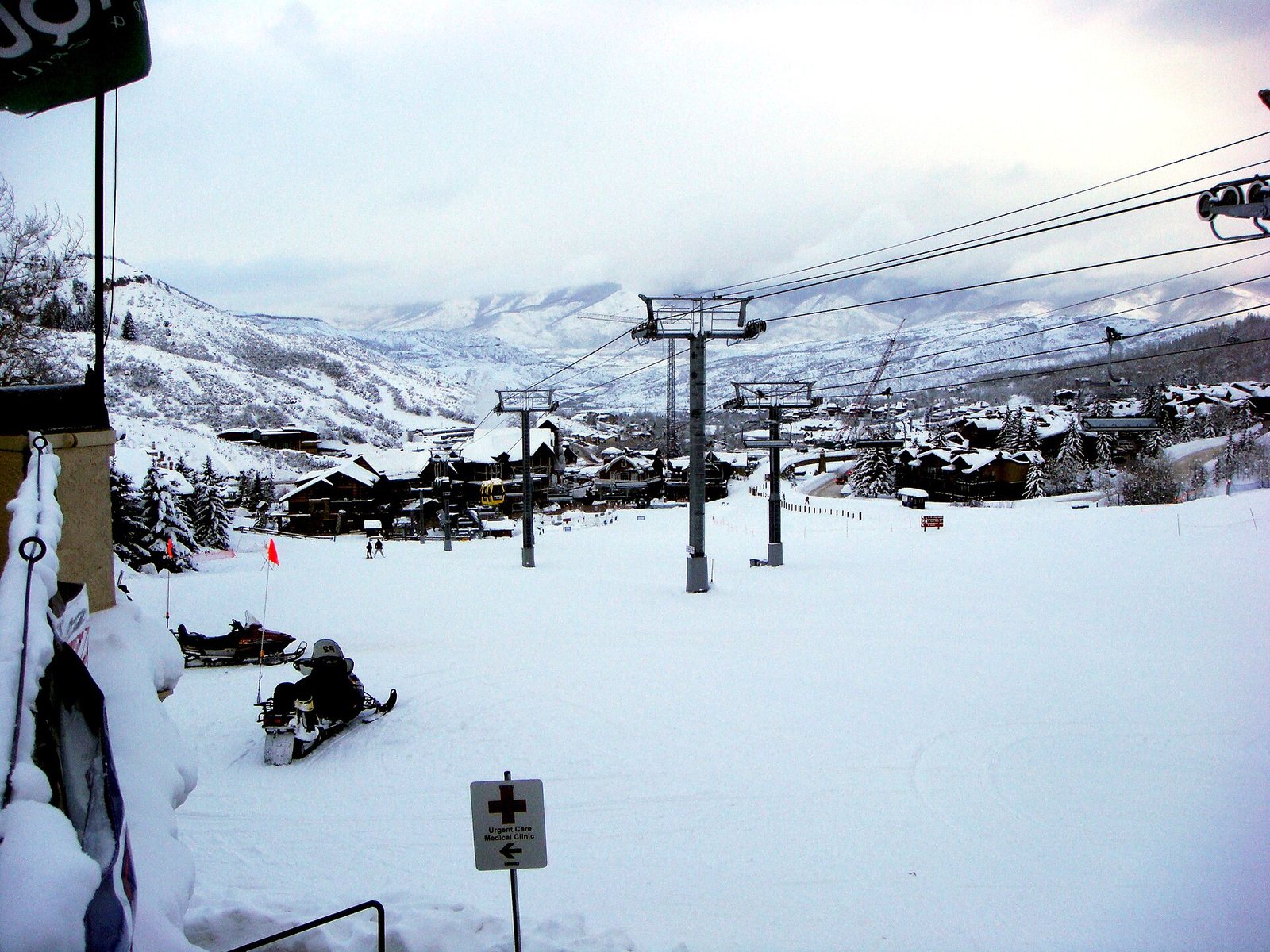 A snow-covered ski slope features multiple ski lift towers, buildings of a village, and distant mountains under an overcast sky, with a snowmobile in the foreground.