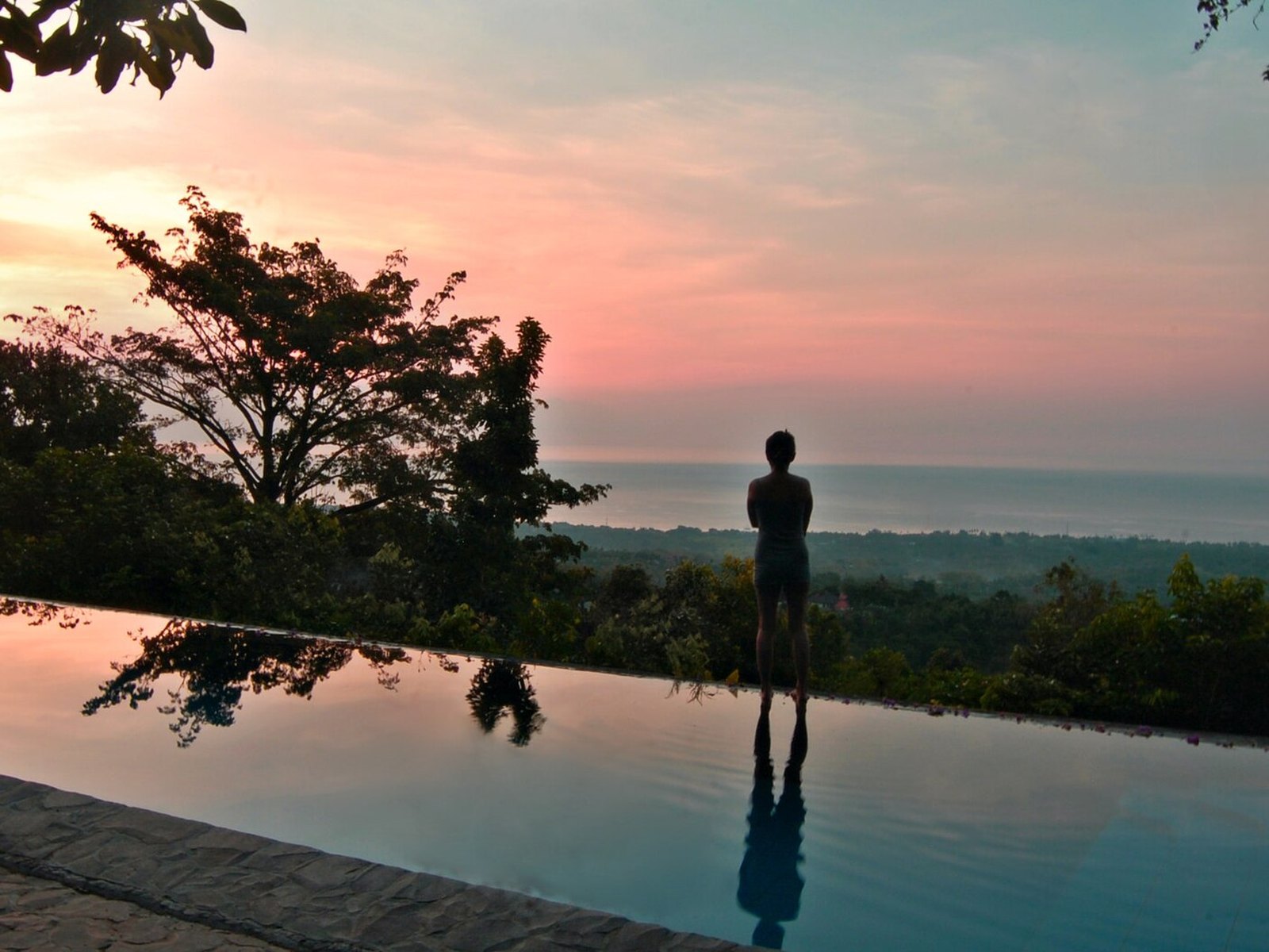 A person standing in silhouette at the edge of an infinity pool at dusk, with trees and a pink sky above the sea