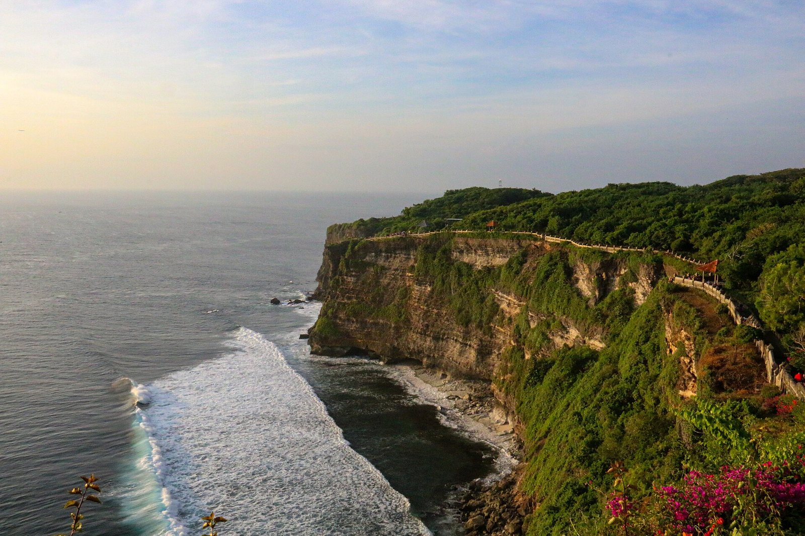 A view shows a vegetated cliff face dropping into the sea, with waves breaking at its base and a path running along the cliff top.