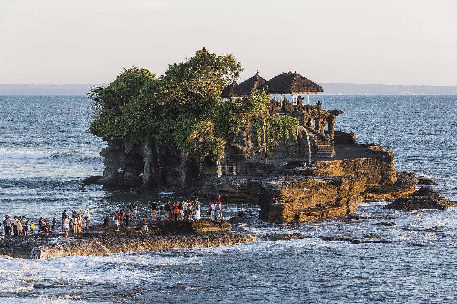 A temple with thatched roofs sits atop a large, vegetated rock formation in the sea, with many people gathered on adjacent lower rocks.