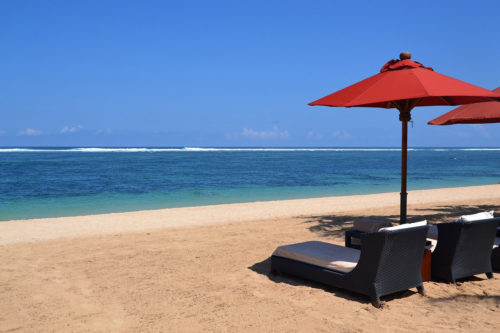 A sandy beach with two sun loungers and a red umbrella in the foreground, facing a calm blue sea under a clear sky.