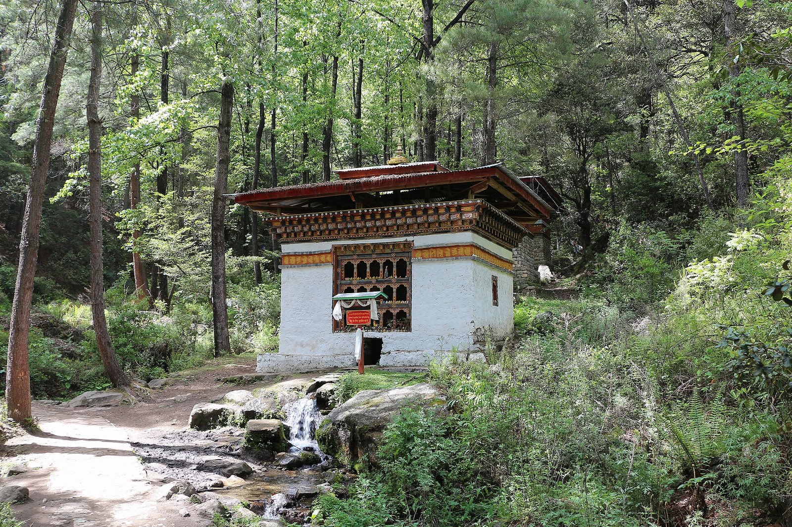 A small white temple with a red roof and ornate windows stands by a stream with a waterfall, next to a dirt path in a dense forest.