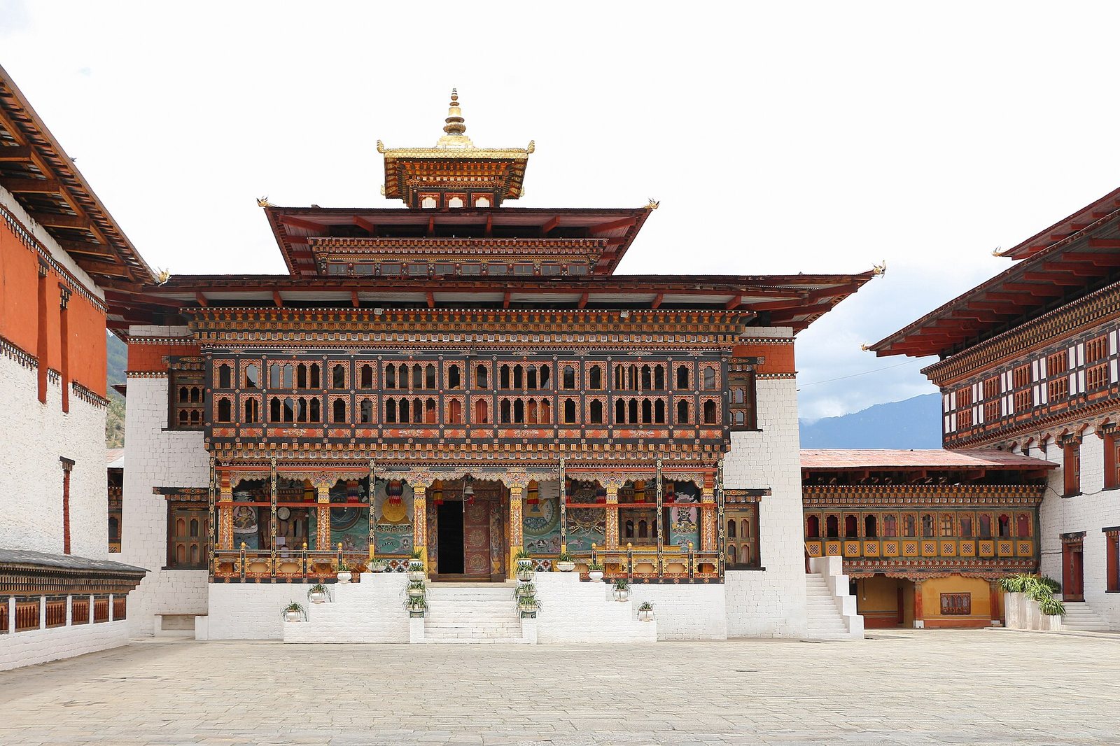 A large, multi-story traditional Bhutanese building with a golden roof ornament, intricate wooden windows, and painted murals, set in a paved courtyard.