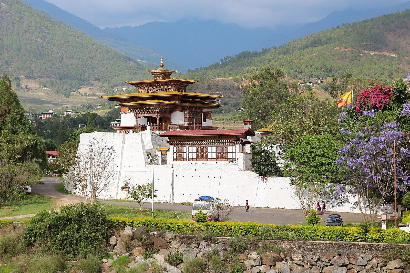 A multi-tiered building with white walls, red roofs, and golden upper sections stands before green mountains. A bus, people, flowering plants, and a Bhutanese flag are in the foreground.