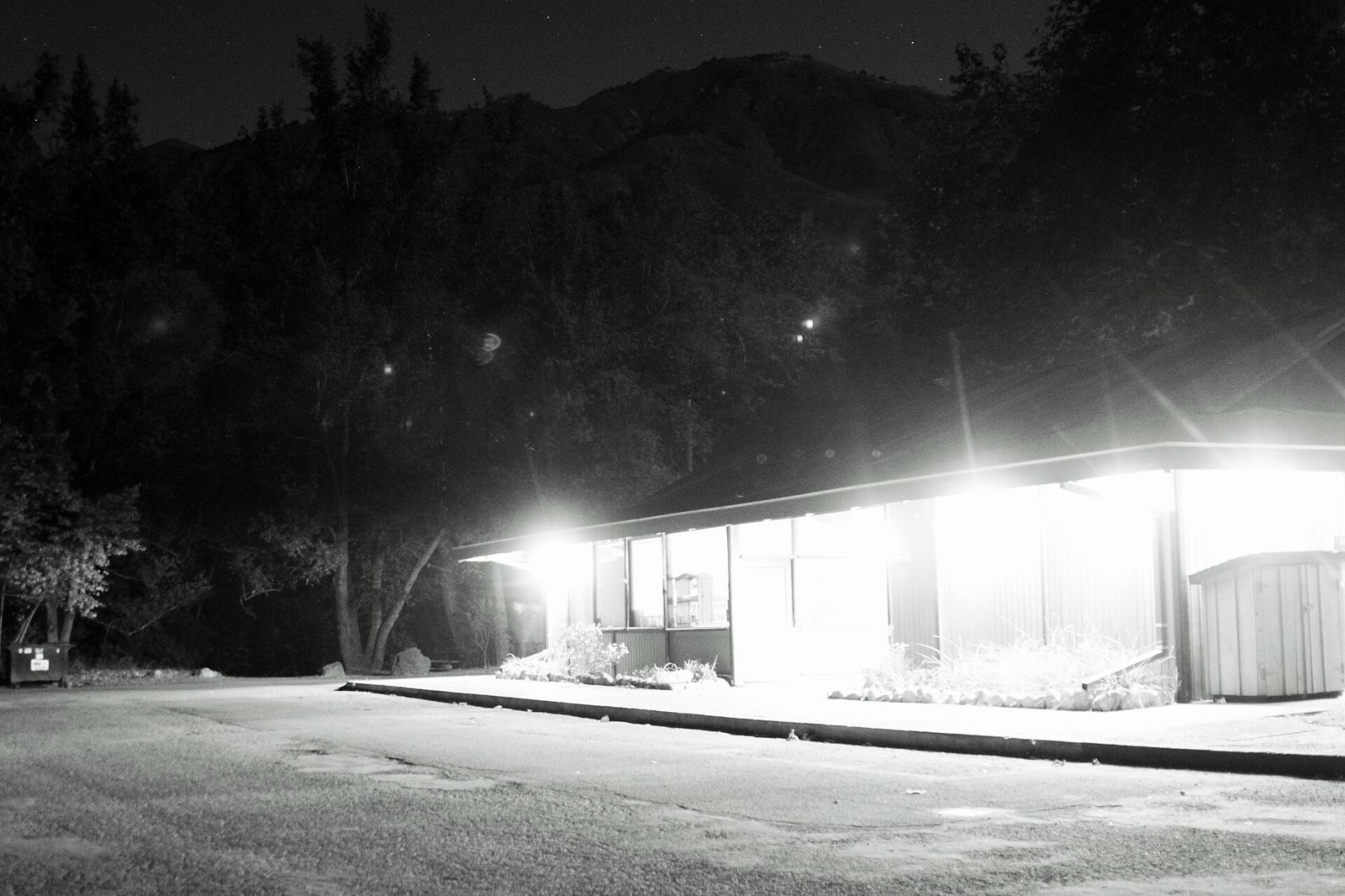 A brightly lit building at night with trees and a dark mountain in the background, and a paved area in the foreground.