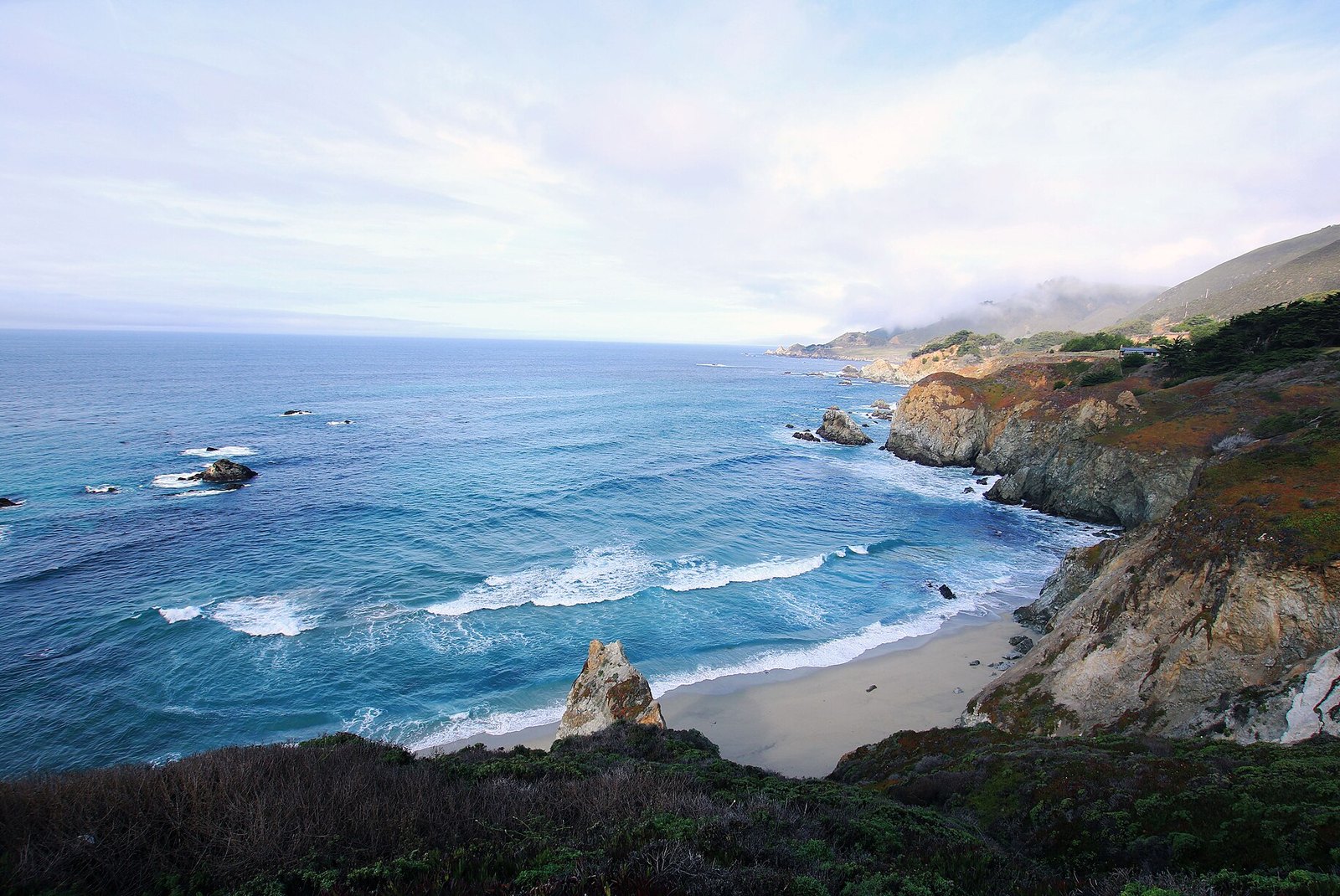 A sandy cove and blue-green surf below a grassy headland on the Big Sur coast, with fog hanging over the hills