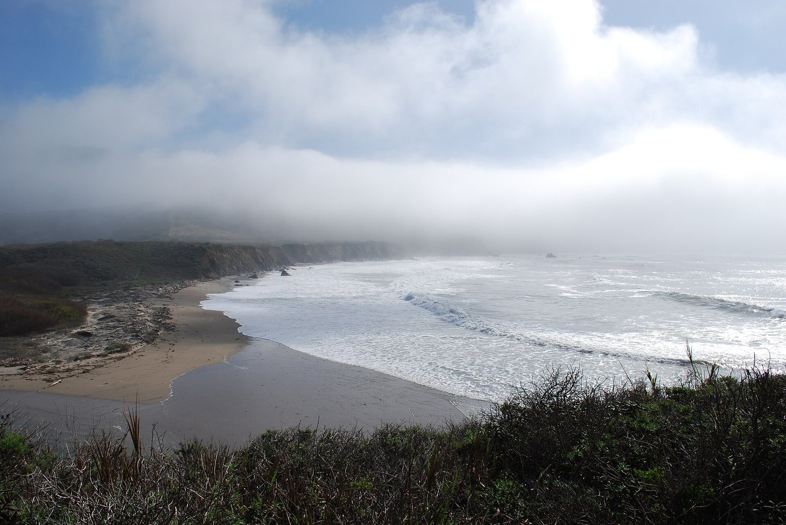 A curving sandy beach meets ocean waves along a rocky coastline with hills in the background, partially obscured by fog, under a cloudy sky.