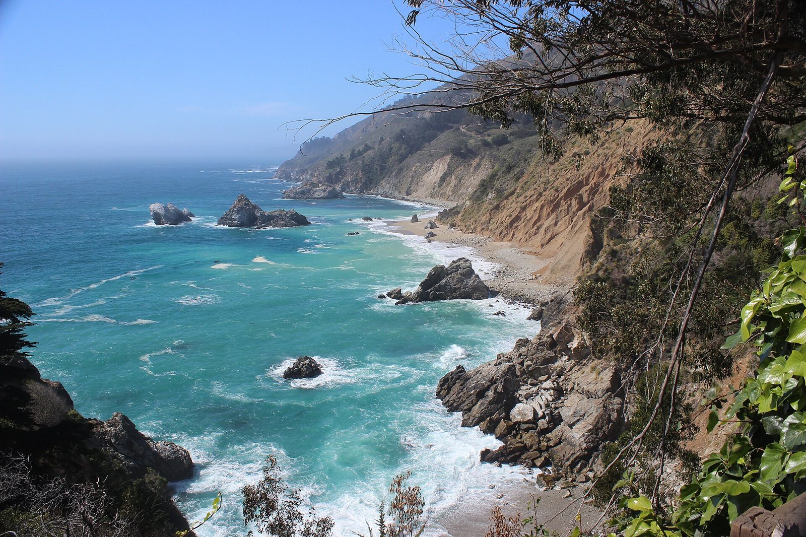 A view of a rocky coastline with turquoise ocean, waves, sea stacks, a narrow pebble beach, and steep, vegetated cliffs under a clear sky.