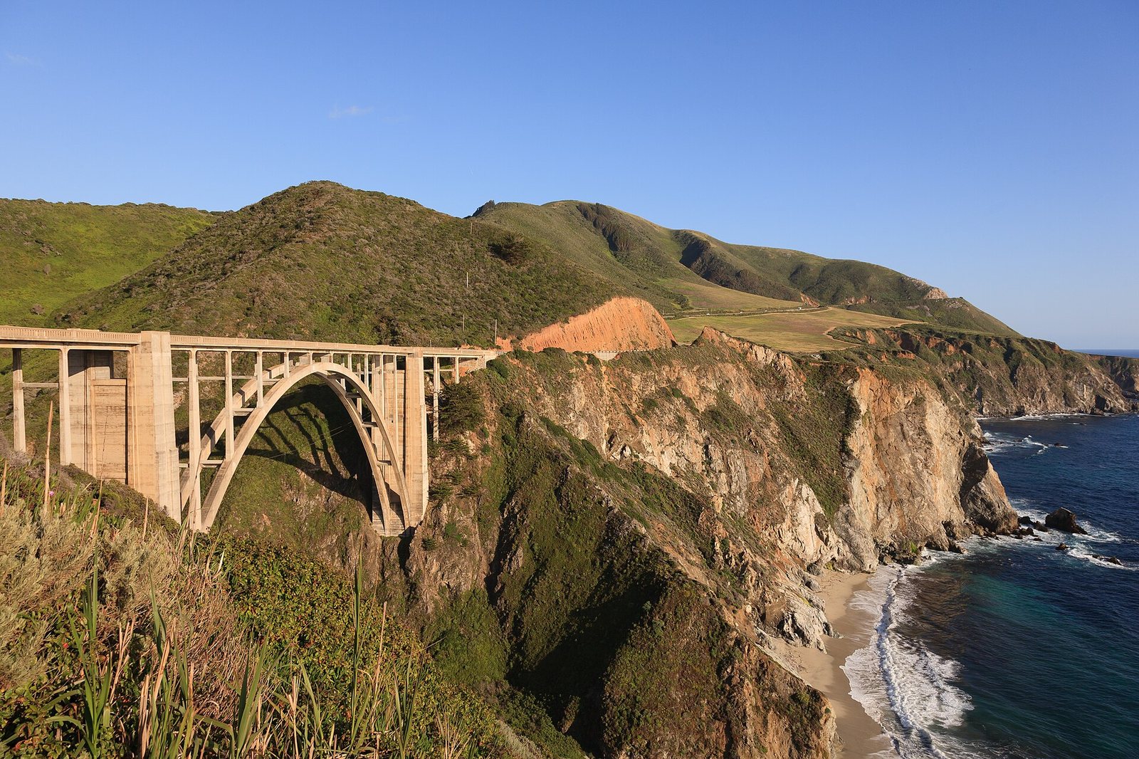 A concrete arch bridge crosses a deep coastal canyon, with green hills in the background and steep cliffs meeting the Pacific Ocean on the right.