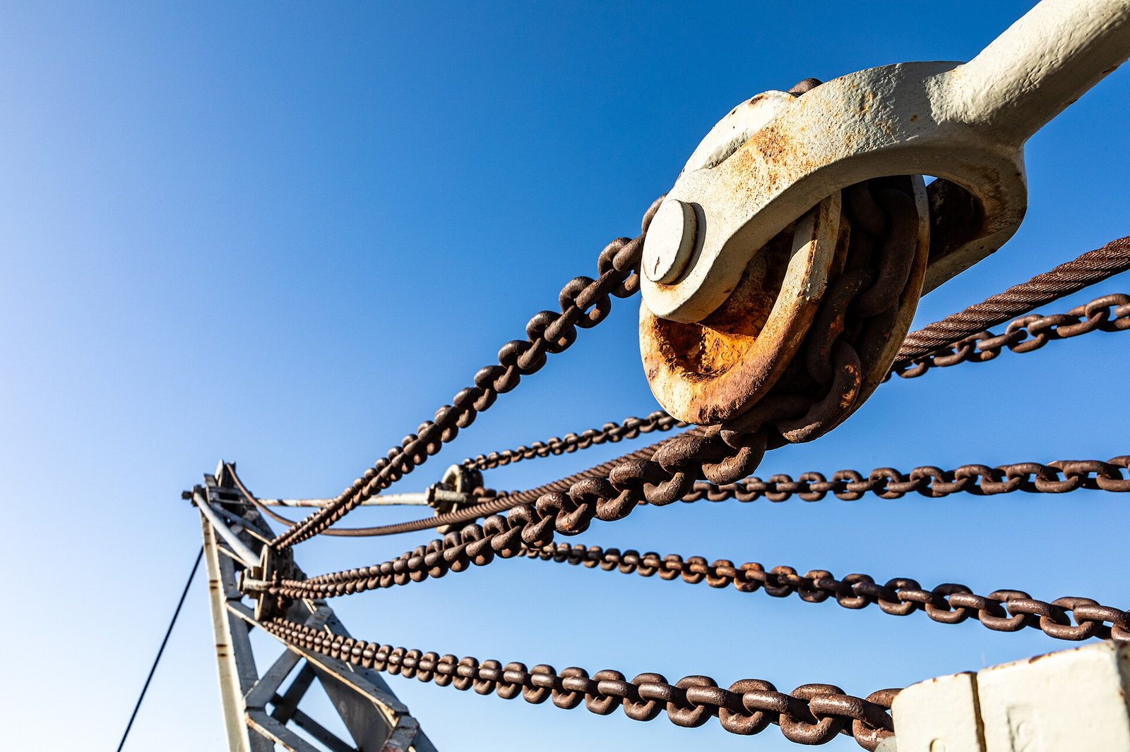 A close-up view of a crane's rusty chains, cables, and a large metal pulley mechanism set against a clear blue sky.