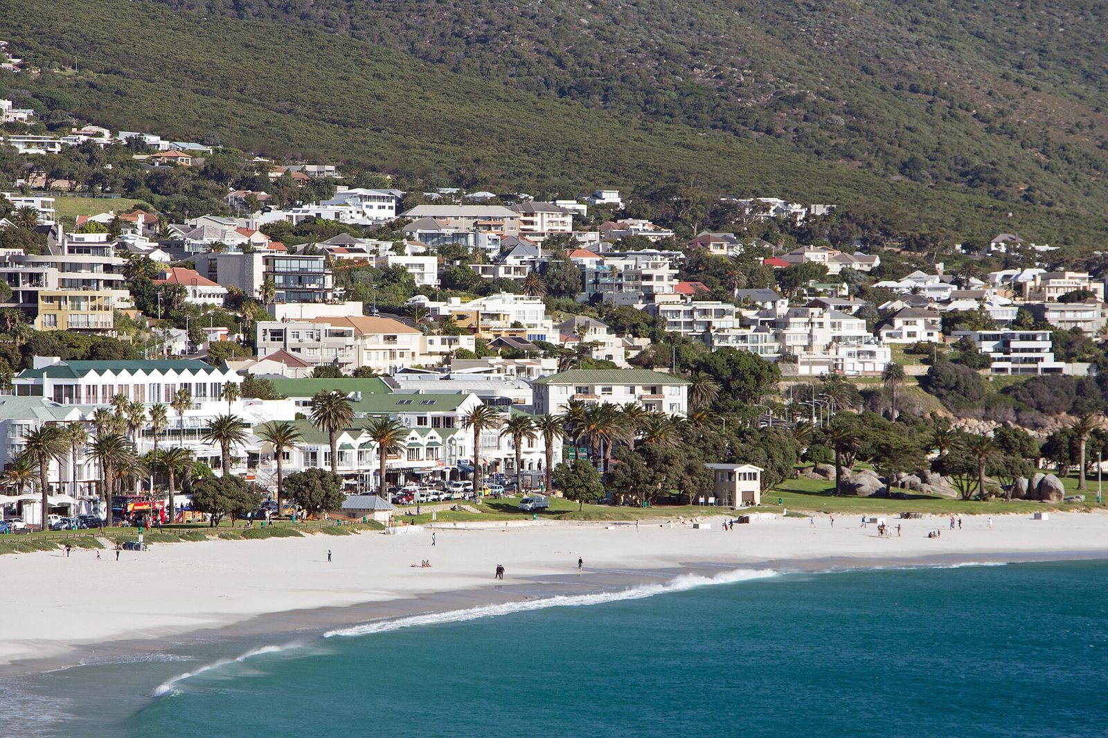 A wide sandy beach with turquoise water and small waves leads to a road lined with buildings and palm trees, backed by houses on a green, tree-covered hillside.