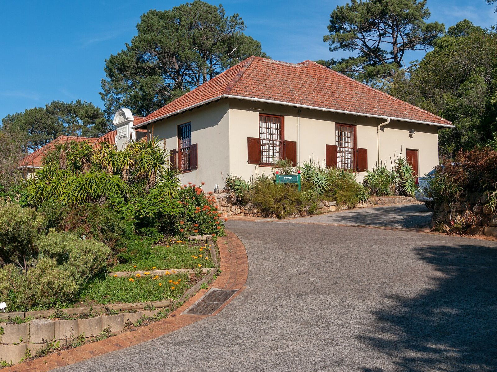 A light-coloured building with a red-tiled roof, brown shutters, and barred windows is surrounded by diverse green plants and orange flowers, with a paved driveway in front and tall trees behind.