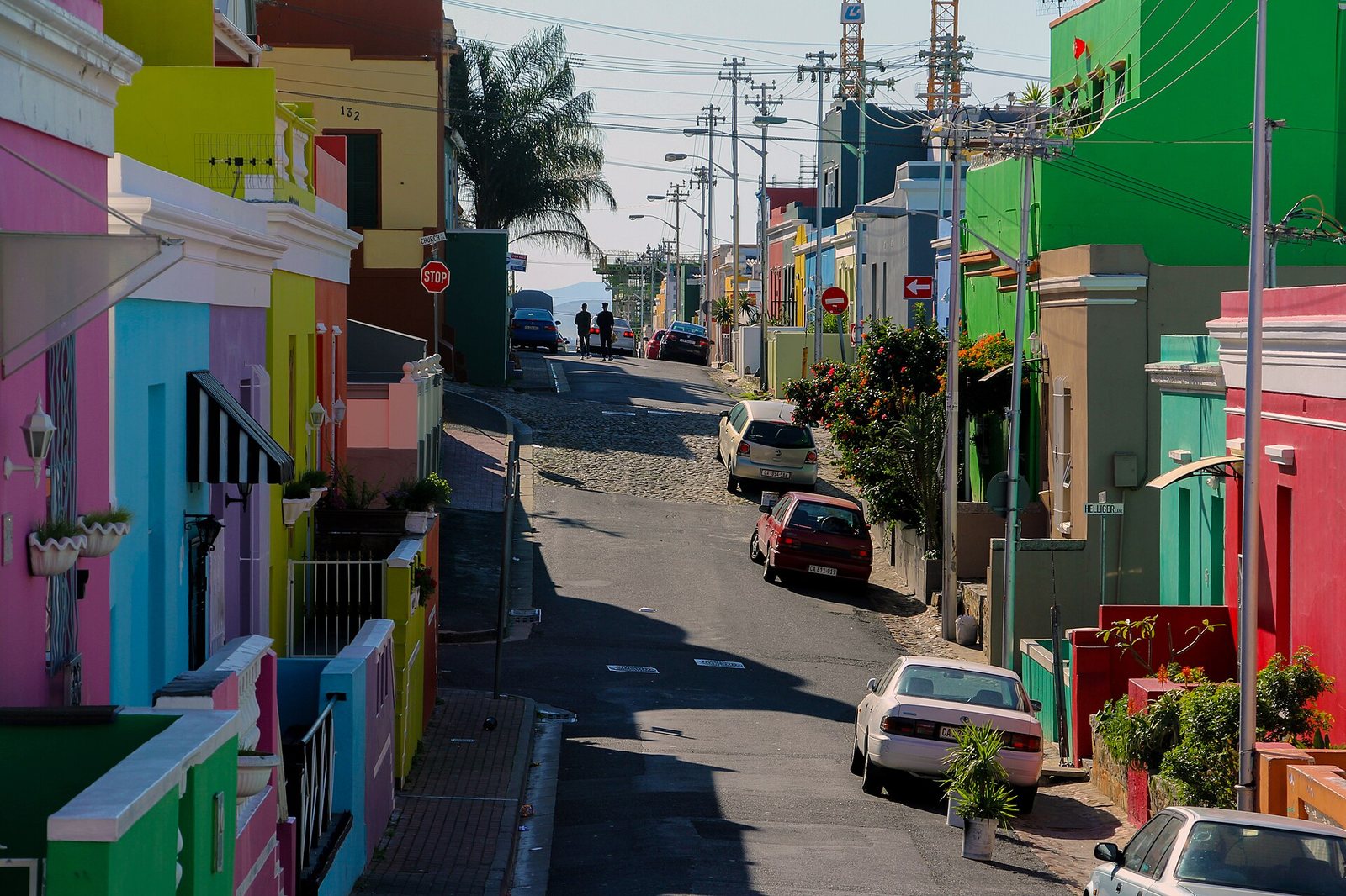 A street slopes downwards, lined with brightly coloured houses, parked cars, and utility poles. In the distance, people stand near a body of water and hills.