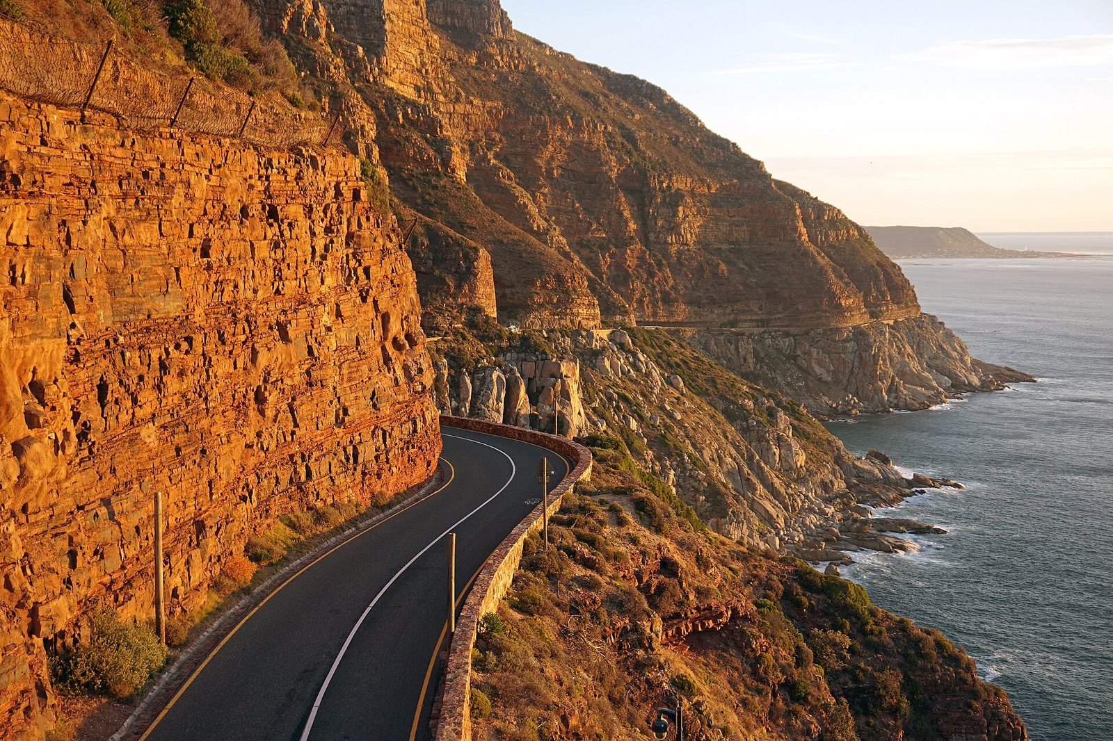 A two-lane road curves along a cliff, with a tall, reddish rock face on the left and the sea with distant land on the right.