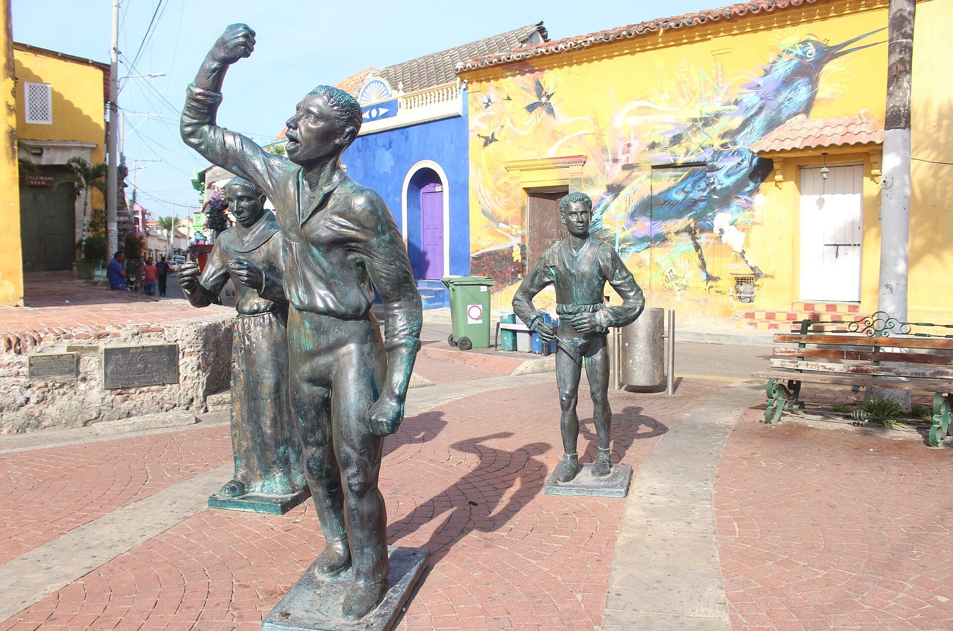 Bronze statues of figures with raised fists and a standing man in a brick-paved plaza in front of yellow and blue painted houses with a mural