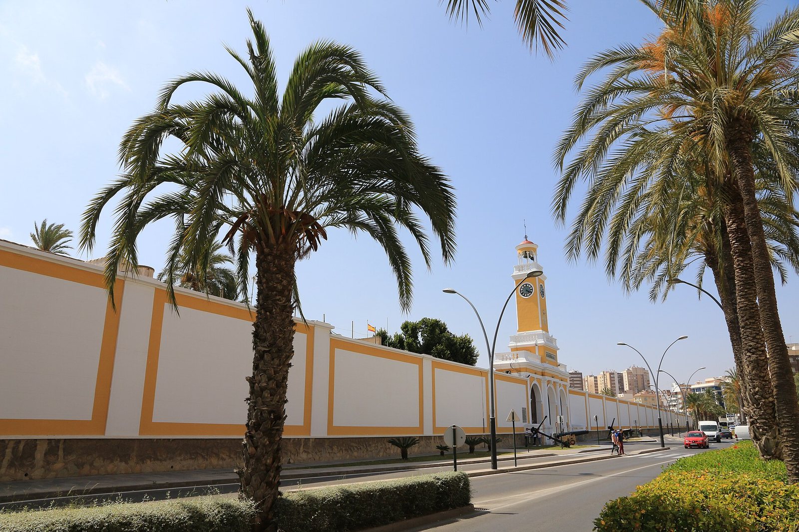 A long white wall with yellow trim, a yellow clock tower, and palm trees line a road with distant buildings under a clear blue sky. An anchor is visible near the tower.