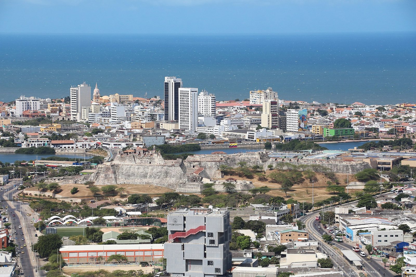 An aerial view of Cartagena city, showing Castle San Felipe de Barajas, modern buildings, roads, and the sea in the background.
