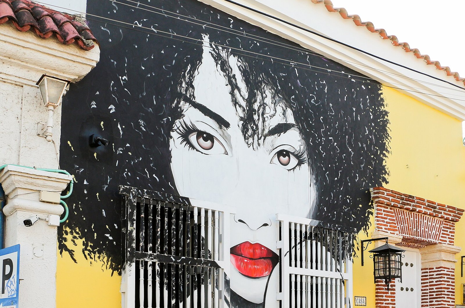 A mural of a woman’s face with dark curly hair and red lips painted on the wall of a yellow colonial house in Getsemaní