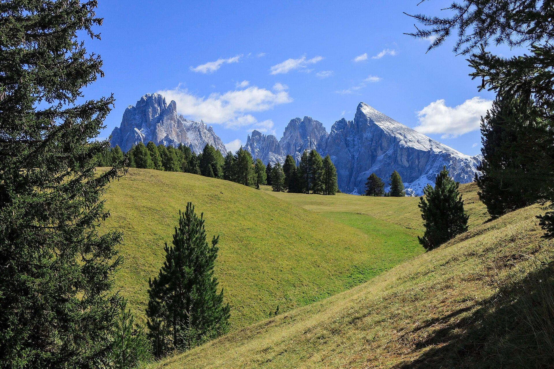 A wide high-altitude meadow rolling toward pale rock towers, with scattered wooden huts and grazing pasture.