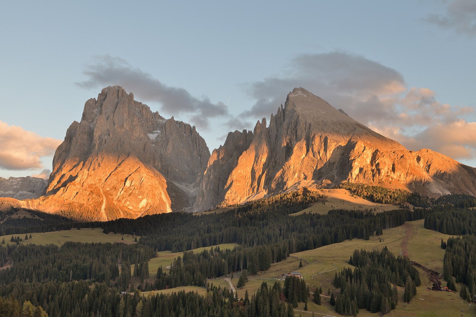 The Langkofel group mountains are seen at sunset, with their peaks illuminated in warm light above a forested and grassy landscape of the Seiser Alm.