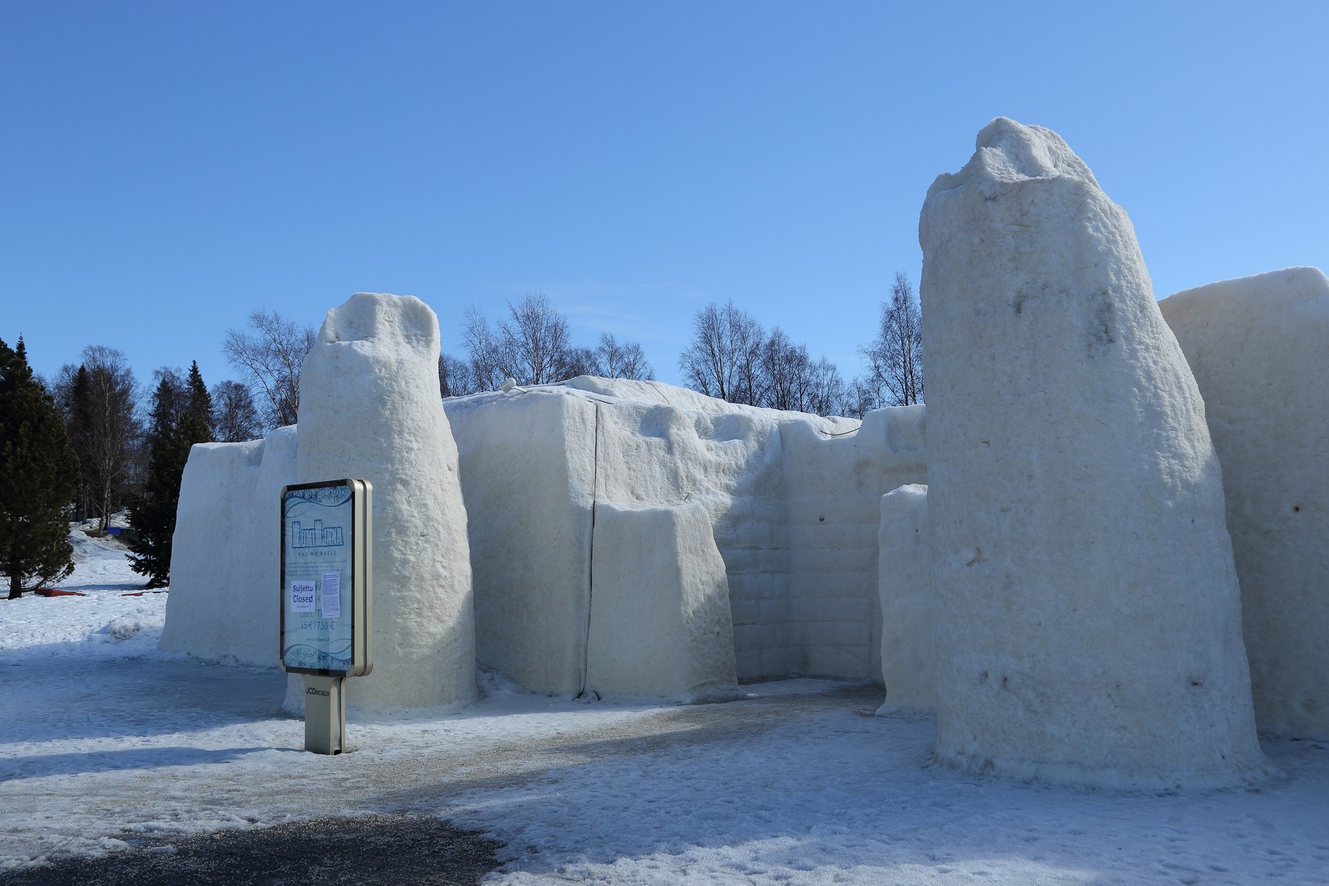Tall walls and towers of white snow and ice at the Kemi SnowCastle under a clear blue winter sky, with pine trees behind