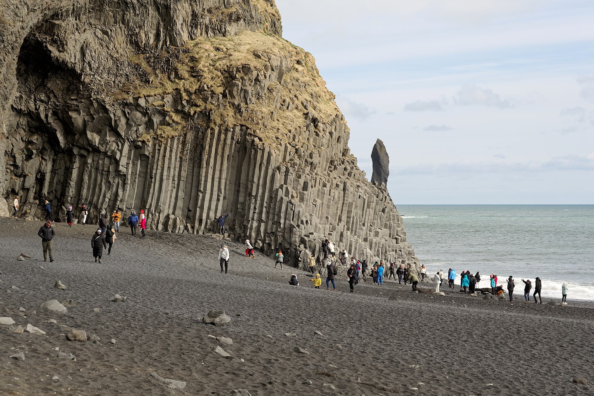 Hexagonal basalt columns forming a cave at the foot of a cliff on a black sand beach, with visitors and the sea beyond